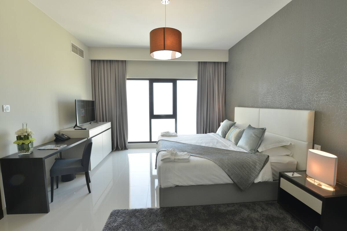 Une chambre d’hôtel moderne équipée d’un lit, d’un bureau et d’une chaise, d’une grande fenêtre et d’un luminaire au plafond.