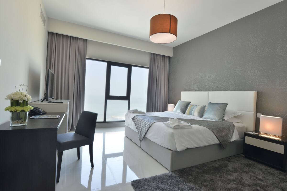 Une chambre d’hôtel moderne dotée d’un grand lit, d’un bureau avec une chaise, d’un téléviseur à écran plat et d’une grande fenêtre avec des rideaux.