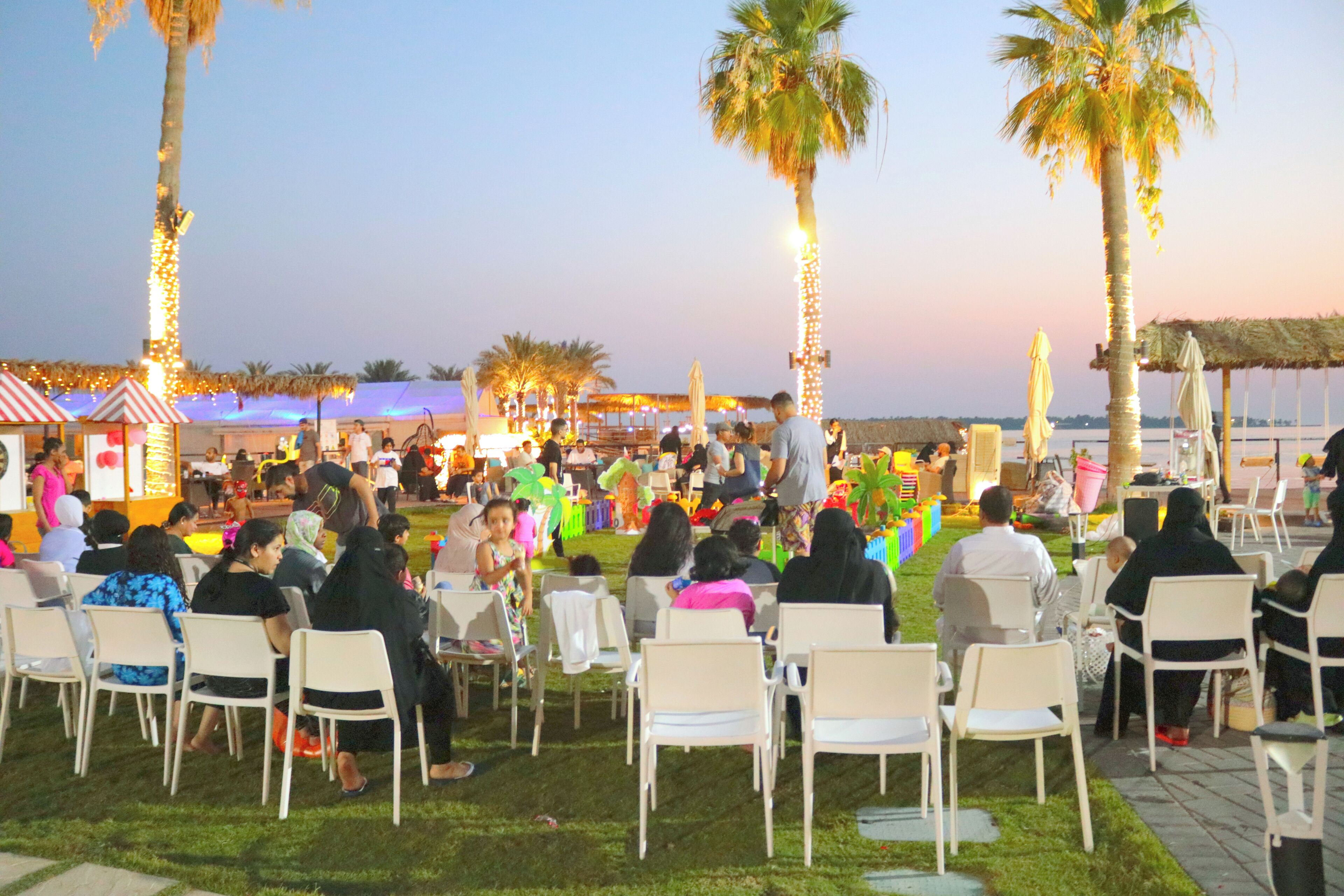 An outdoor event with attendees seated on white chairs, palm trees, and string lights in the background.