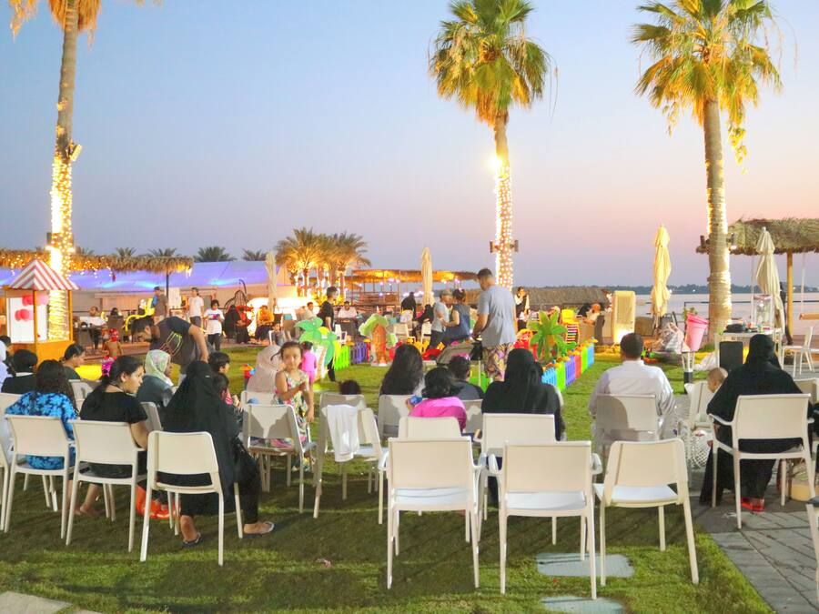 An outdoor event with attendees seated on white chairs, palm trees, and string lights in the background.
