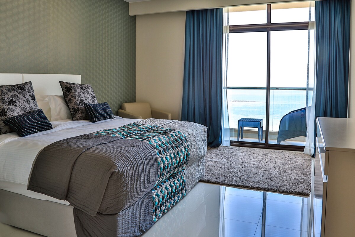 Habitación de hotel con una cama grande, una silla, una mesita y vista al mar por la ventana.