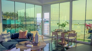 Un salon moderne avec un canapé, une table basse et un coin repas, de grandes fenêtres en verre et une vue sur une piscine et des palmiers.