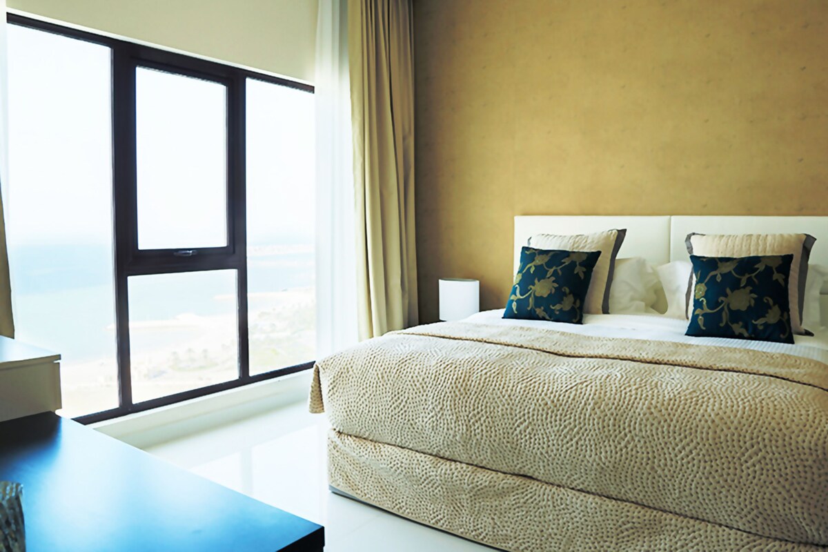 Una habitación de hotel con un ventanal grande, una cama con una colcha color beige y dos almohadas decorativas.
