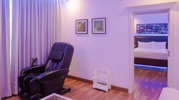 A hotel room with a massage chair, a bed with a blue headboard, and two framed pictures on the wall.