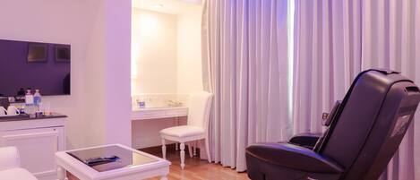 A modern hotel room with a massage chair, a small table with a book, a white chair, a vanity with a mirror, and a large window with curtains.