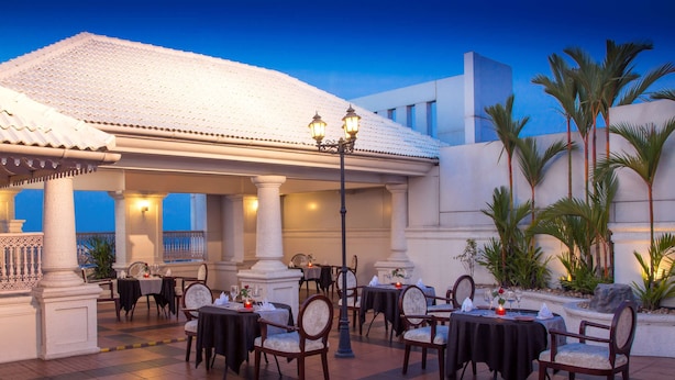 An outdoor dining area with tables and chairs, a covered section with columns, and a view of the sky. - Radisson Blu Kochi (Kanayannur)