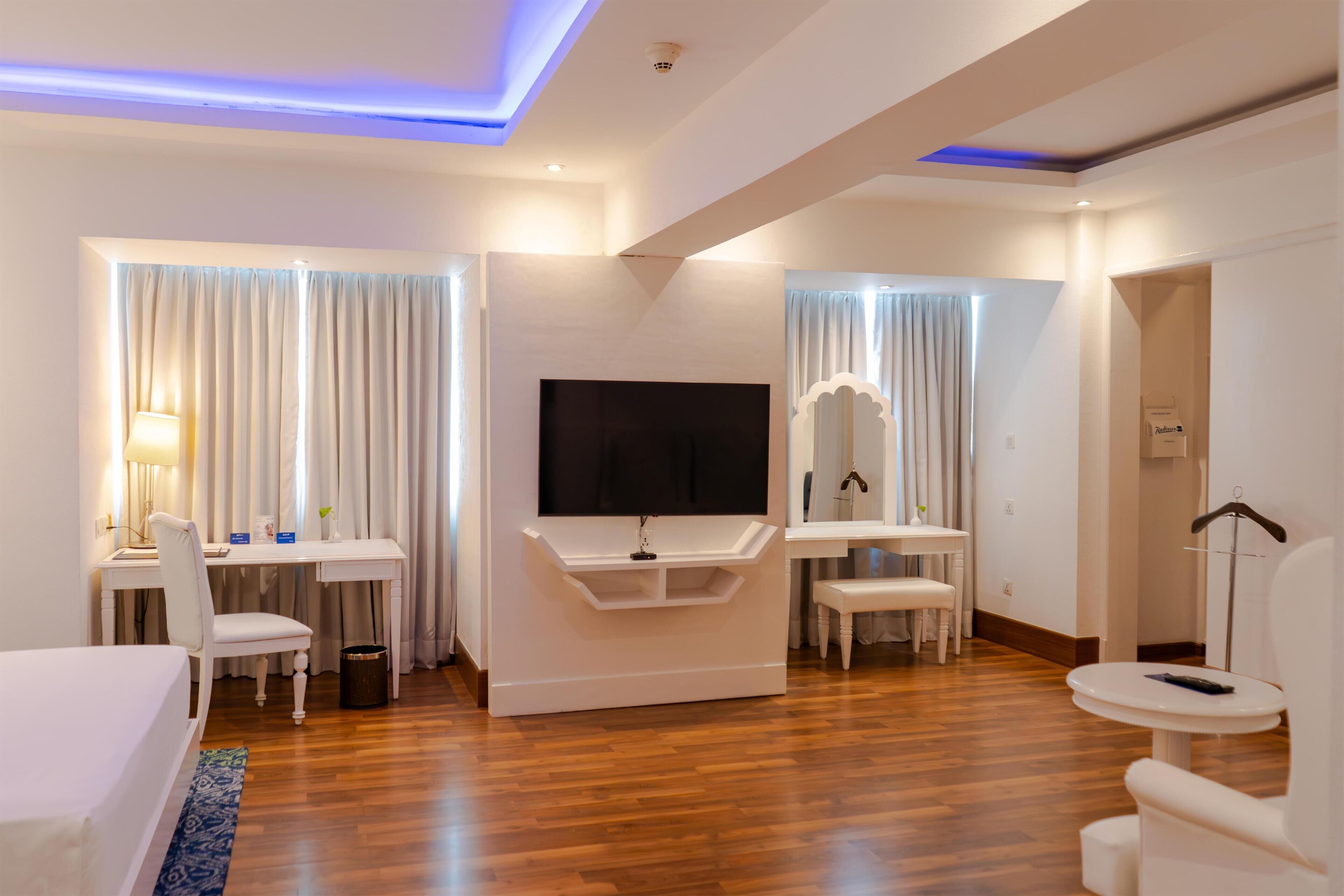 Une chambre d’hôtel équipée d’une télévision à écran plat, d’un bureau avec une chaise, d’une petite table, d’un canapé et d’un porte-manteau mural.