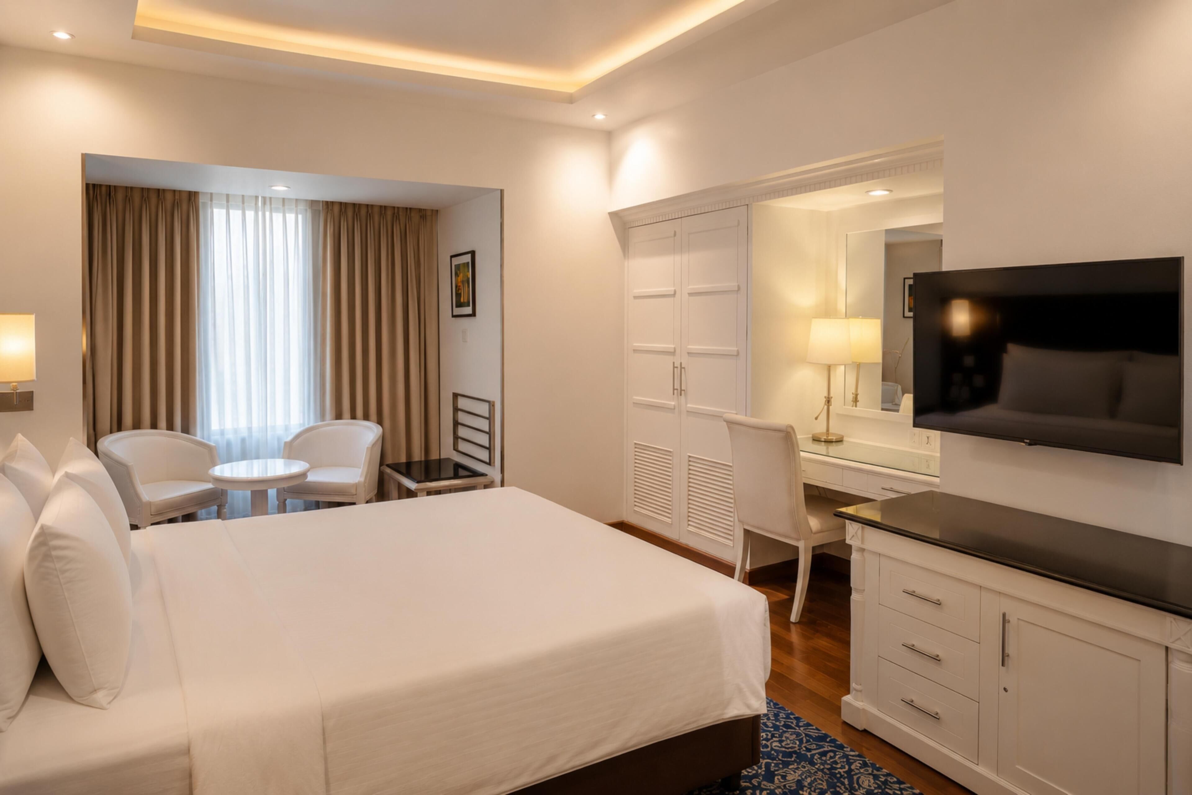 A hotel room with a bed, two chairs, a desk, a TV, and a wardrobe.