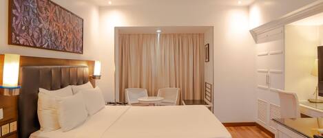 A hotel room with a bed, bedside lamps, a desk with a telephone, a chair, a small table, a mirror, and a painting on the wall.