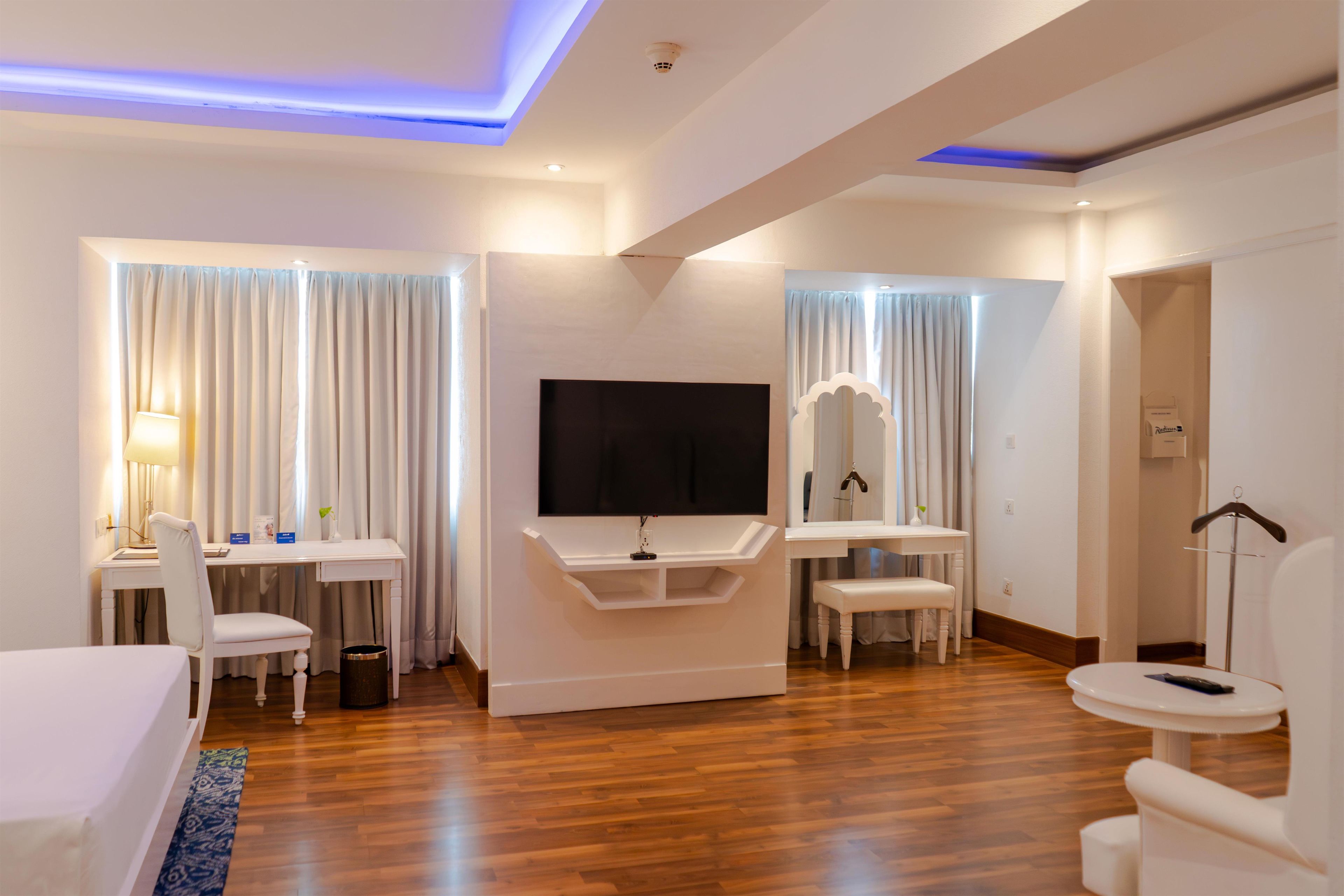 A hotel room with a flat-screen TV, a desk with a chair, a bedside table, a mirror, and a hanging coat rack.