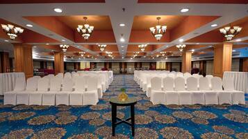 A banquet hall with rows of white chairs, a blue carpet with a floral pattern, and chandeliers on the ceiling.