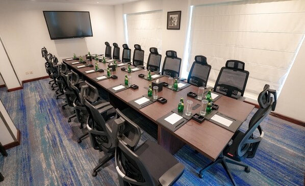 Un salón de conferencias con una larga mesa, varias sillas, un televisor de pantalla plana y una ventana con persianas.