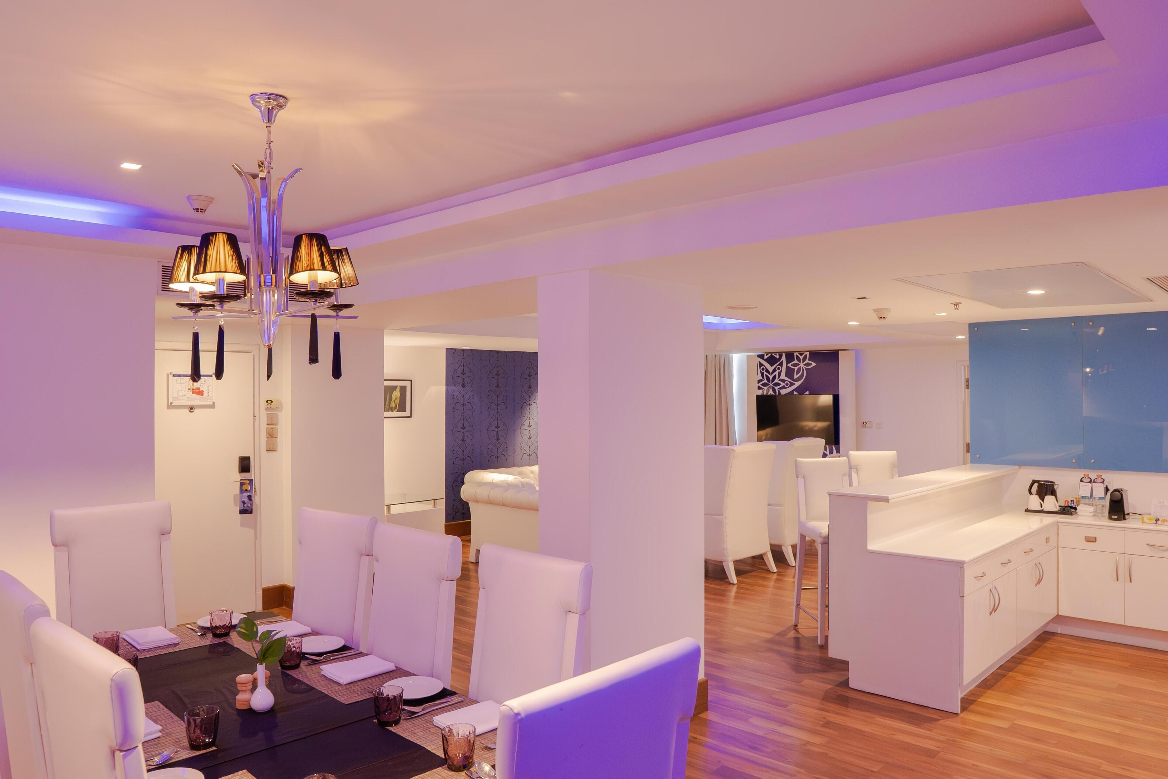 Uma sala de jantar moderna com mobiliário branco, um candelabro e uma cozinha ao fundo.