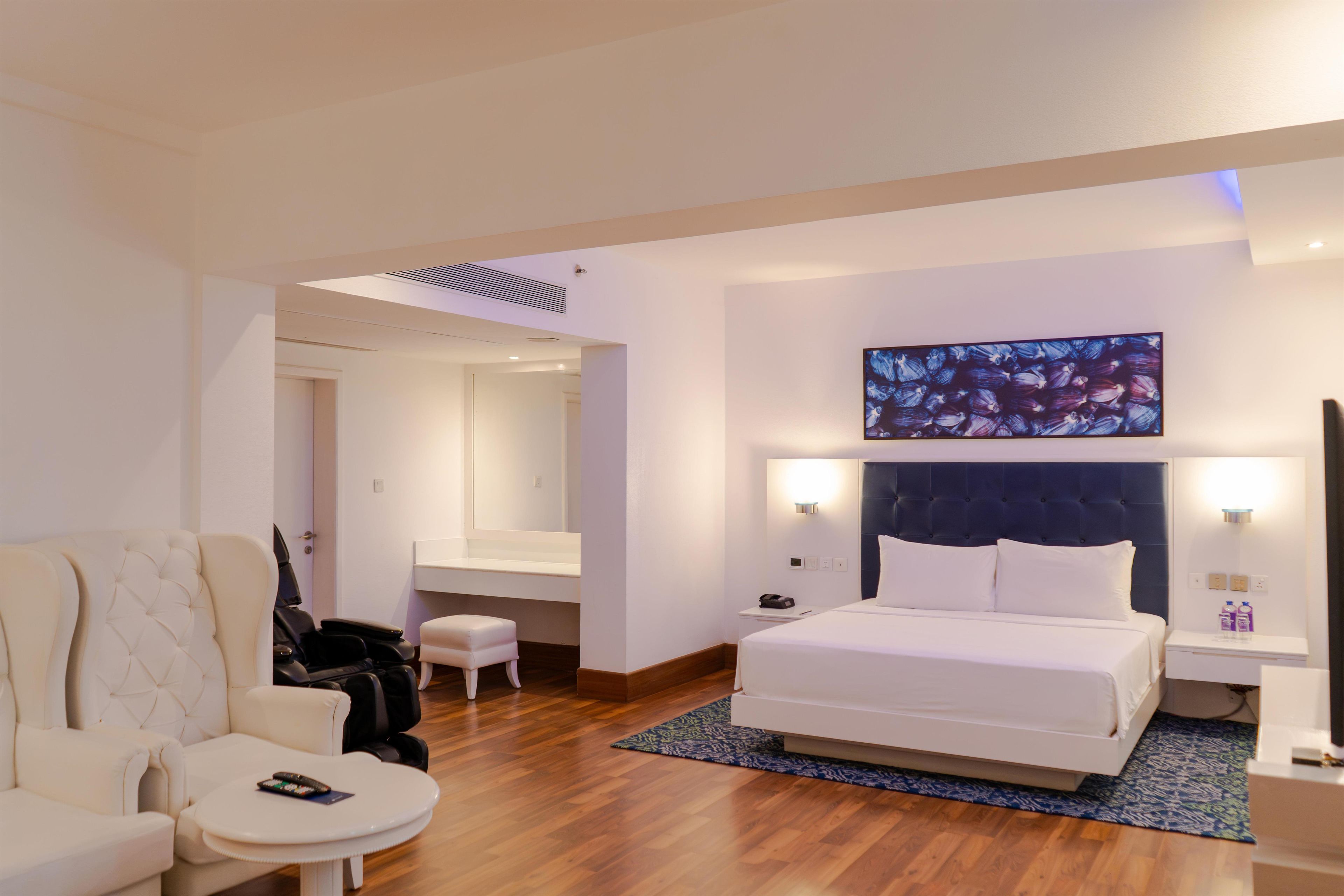 A hotel room with a bed, a seating area with a chair and a small table, a large mirror, and a wall-mounted artwork.