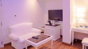 A hotel room with a white armchair, a small table with a remote control, a minibar, a desk with a lamp, and a mirror.