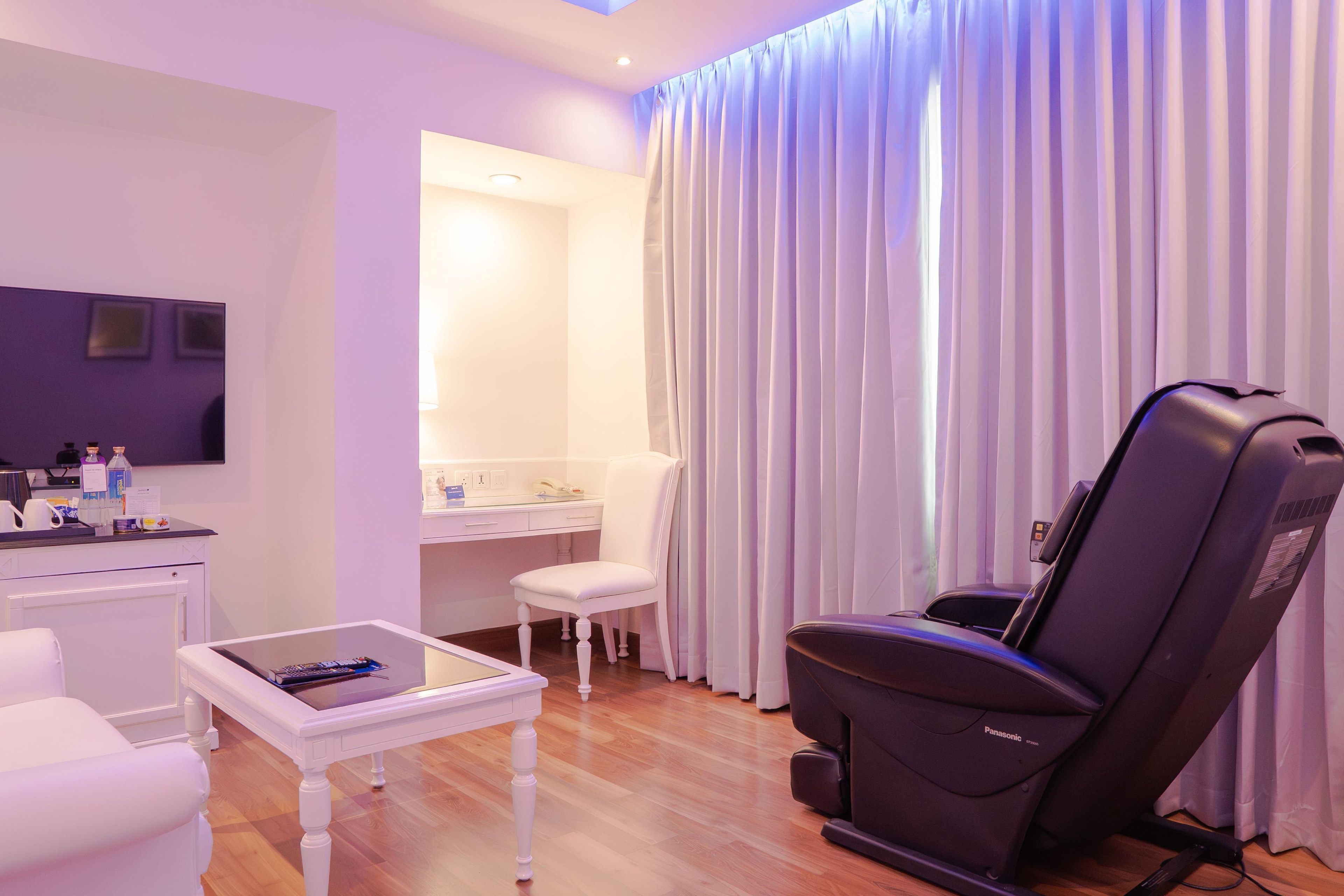 A hotel room with a massage chair, a small table with a remote control, a white chair, and a vanity with a mirror.