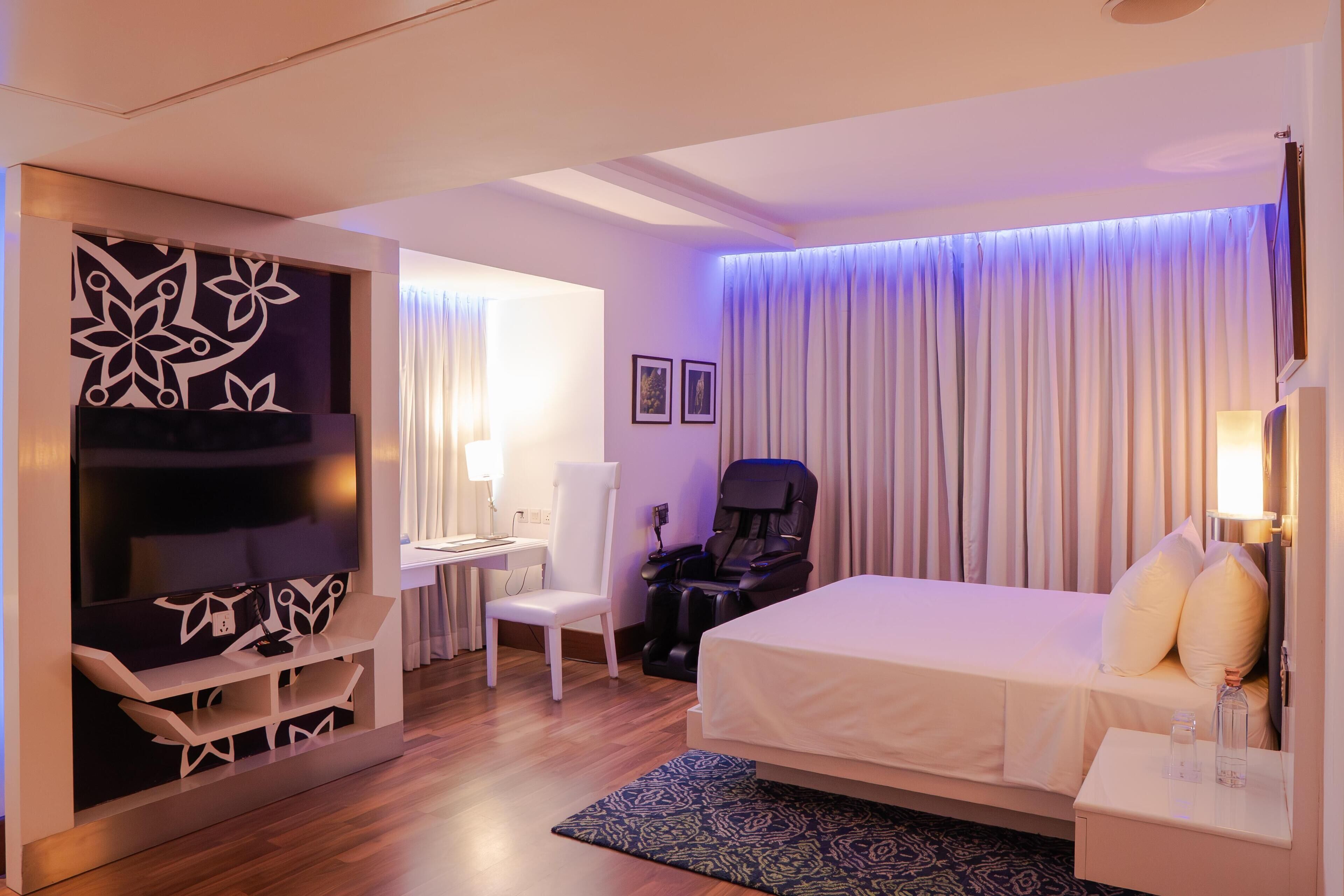 A hotel room with a bed, a desk, a chair, a massage chair, and a TV.