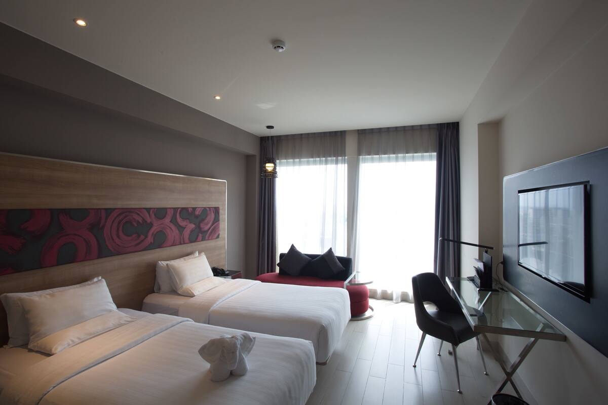 A hotel room with two beds, a desk, a chair, and a large window with curtains.