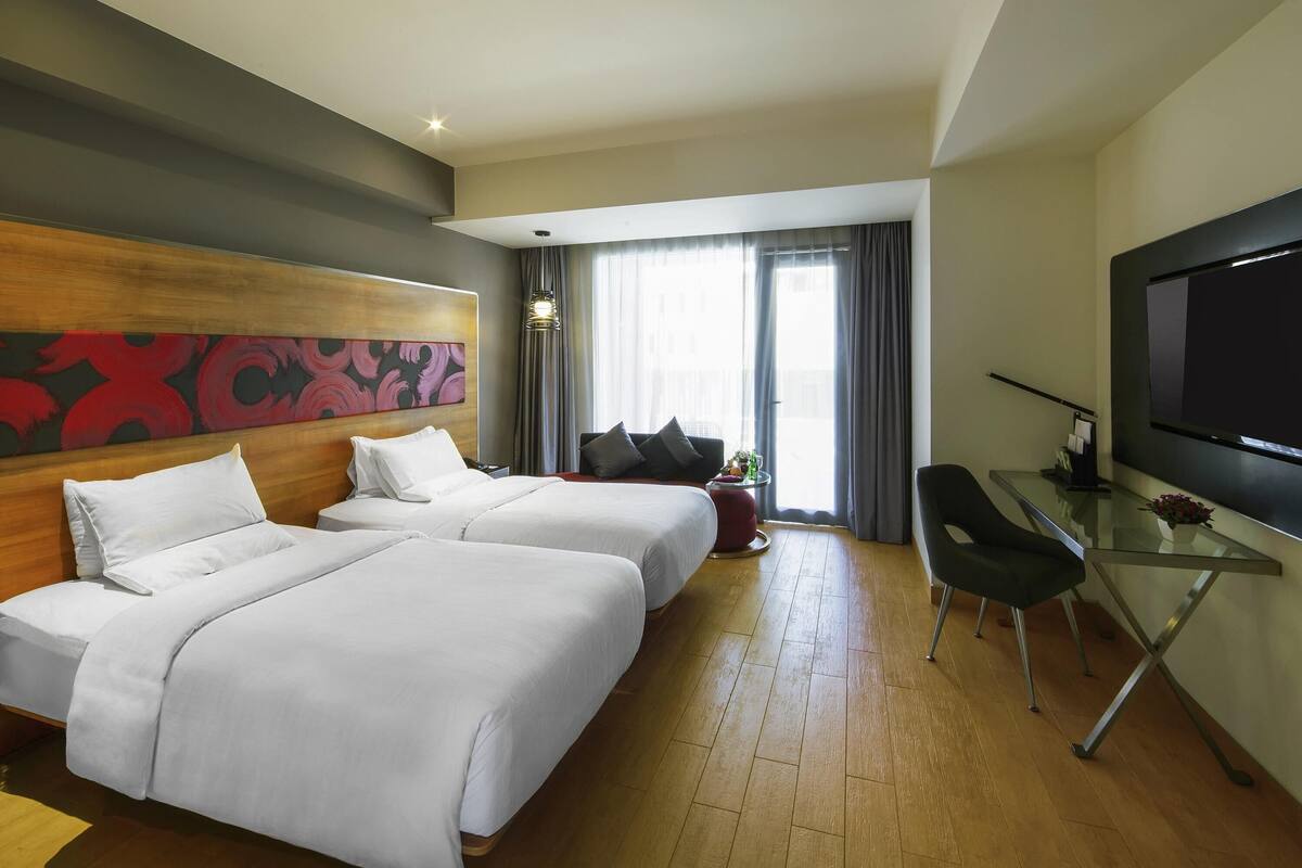 A hotel room with two beds, a TV, a desk, and a large window with curtains.