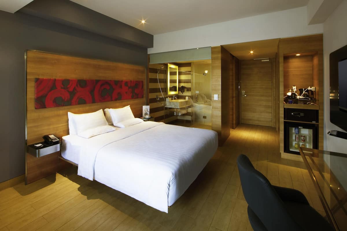 A modern hotel room with a large bed, a desk with a chair, a walk-in closet, and a bathroom with a glass shower enclosure.
