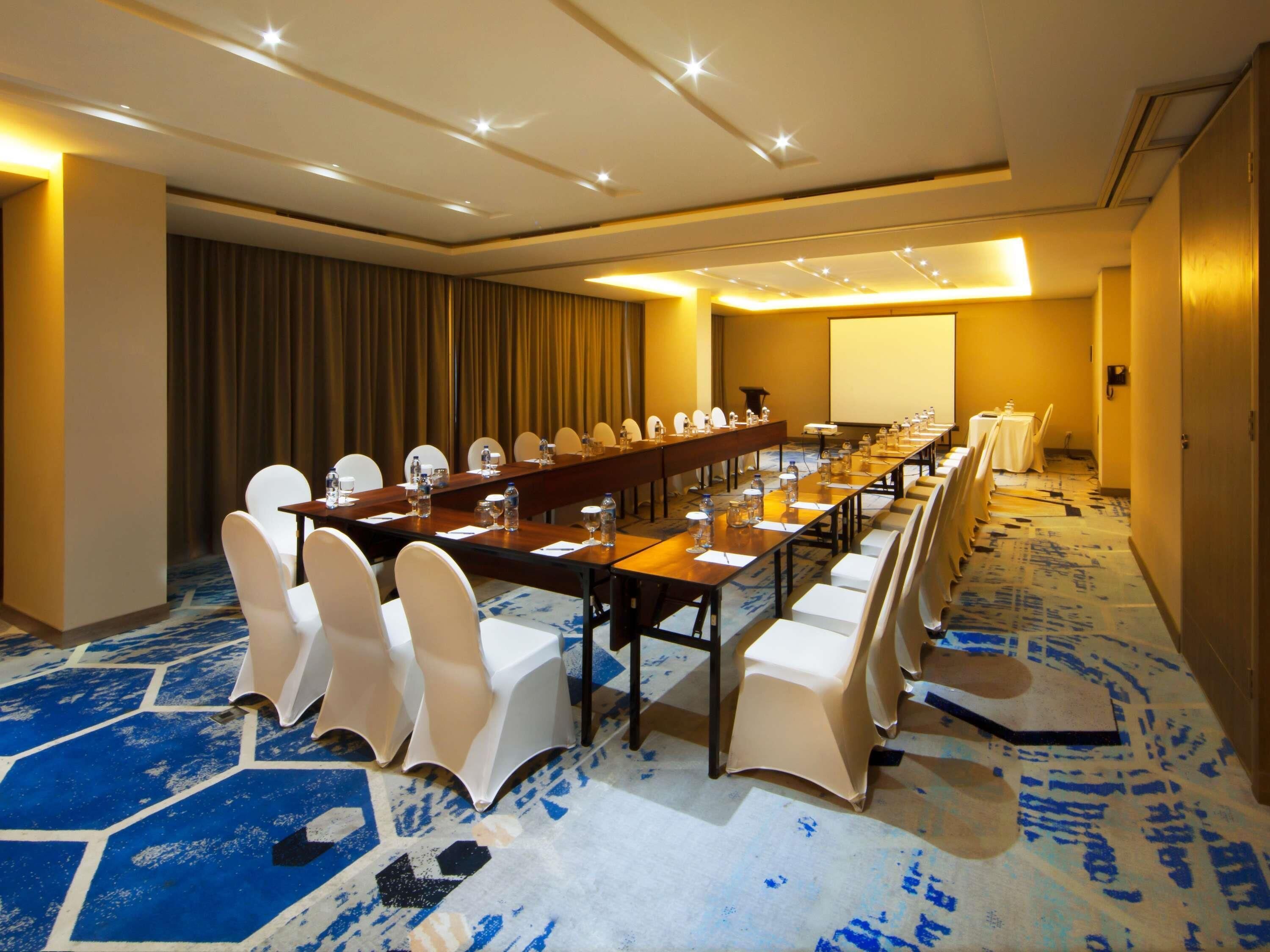 A conference room with a long table, white chairs, and a projector screen.