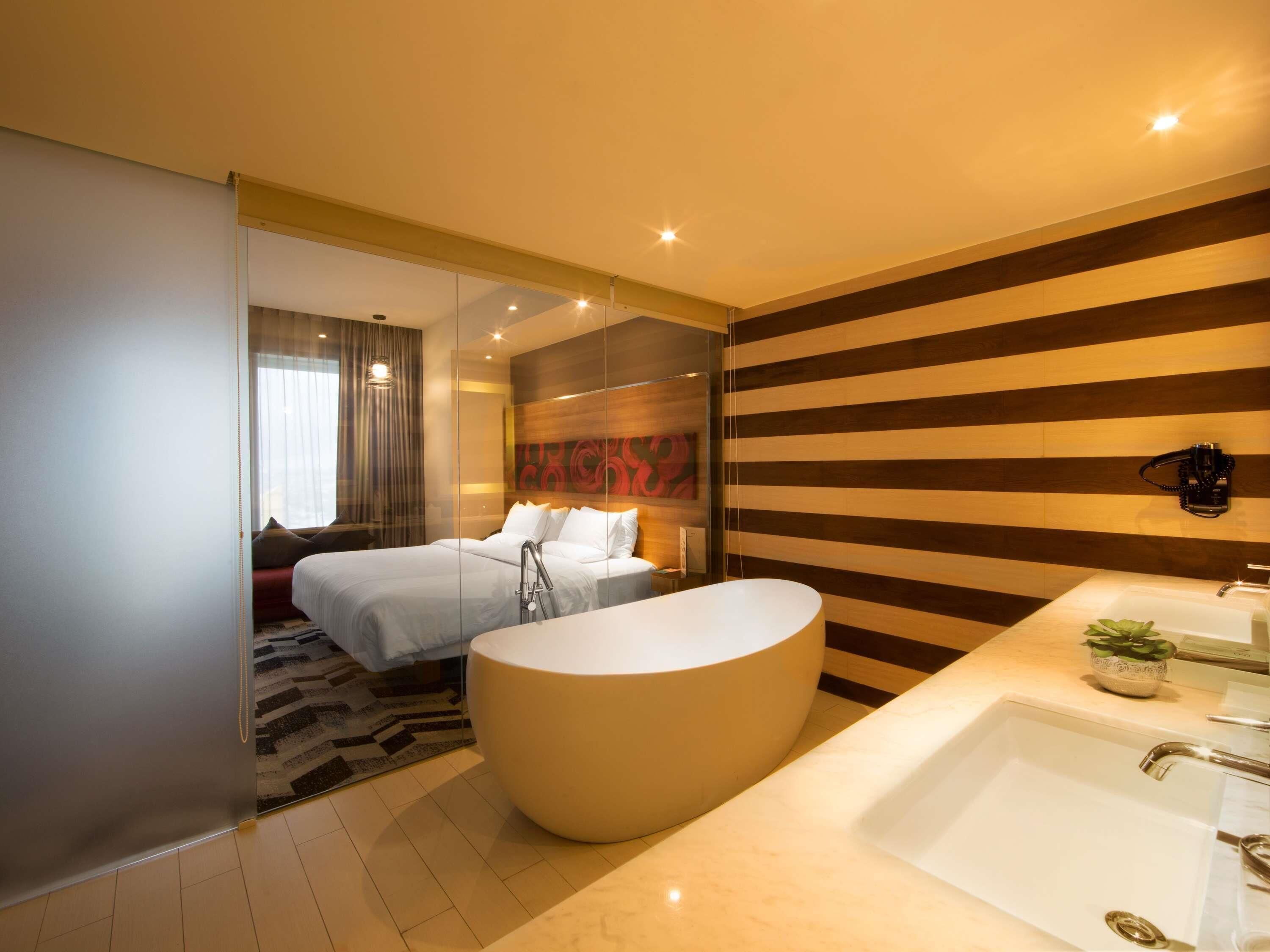 Habitación de hotel moderna con una cama grande, un separador de vidrio, una bañera amplia y un baño moderno con lavamanos y espejo.