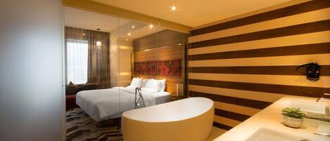 Habitación de hotel moderna con una cama grande, un separador de vidrio, una bañera amplia y un baño moderno con lavamanos y espejo.