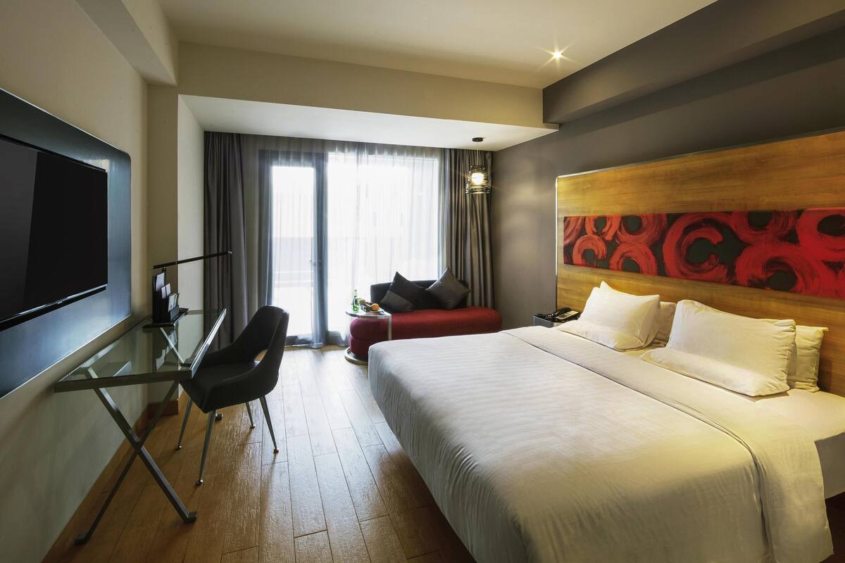A hotel room with a bed, a desk with a chair, a TV, a window with curtains, and a wall with a red abstract design.