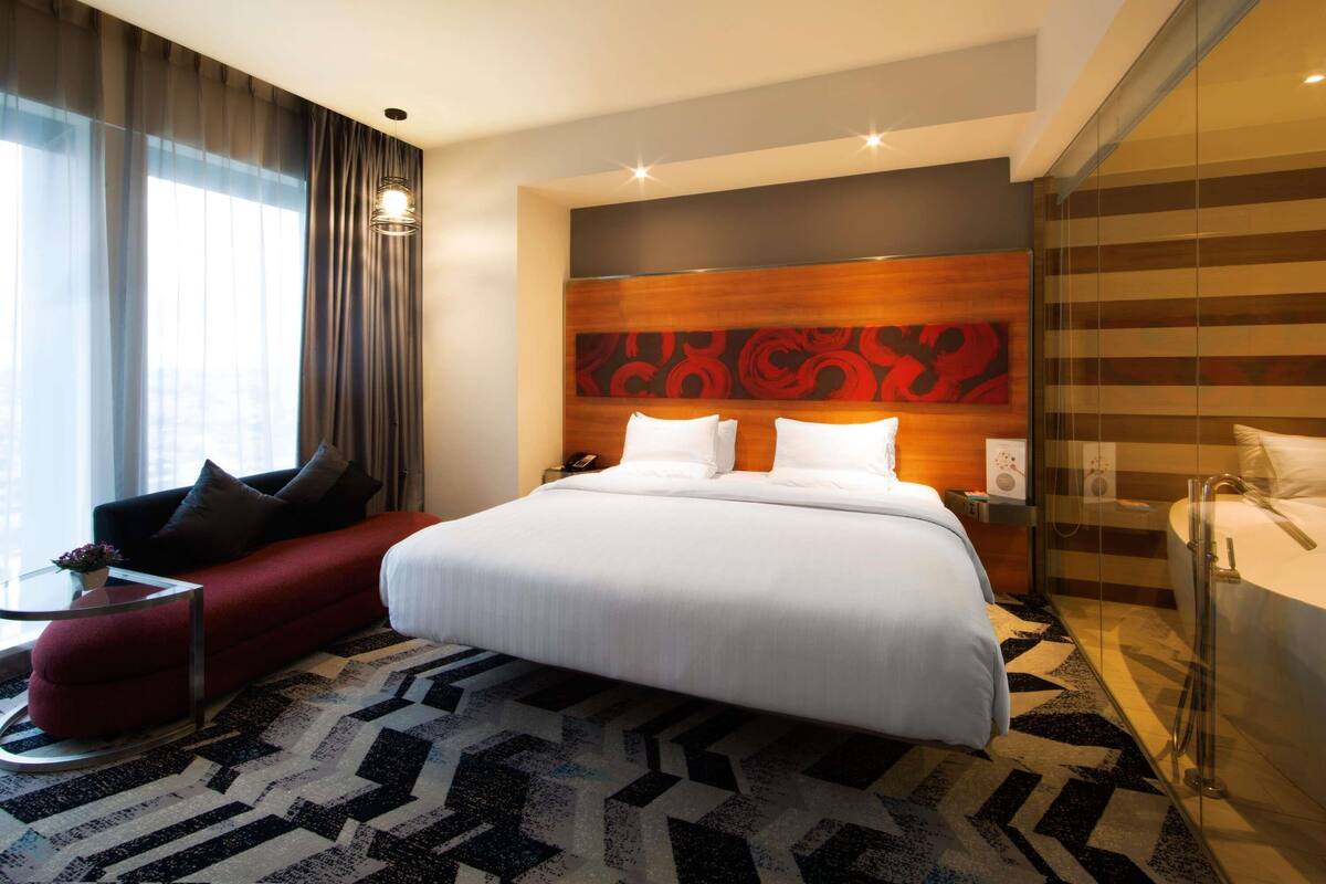 A hotel room with a large bed, a red sofa, a glass partition, and a view of the city.