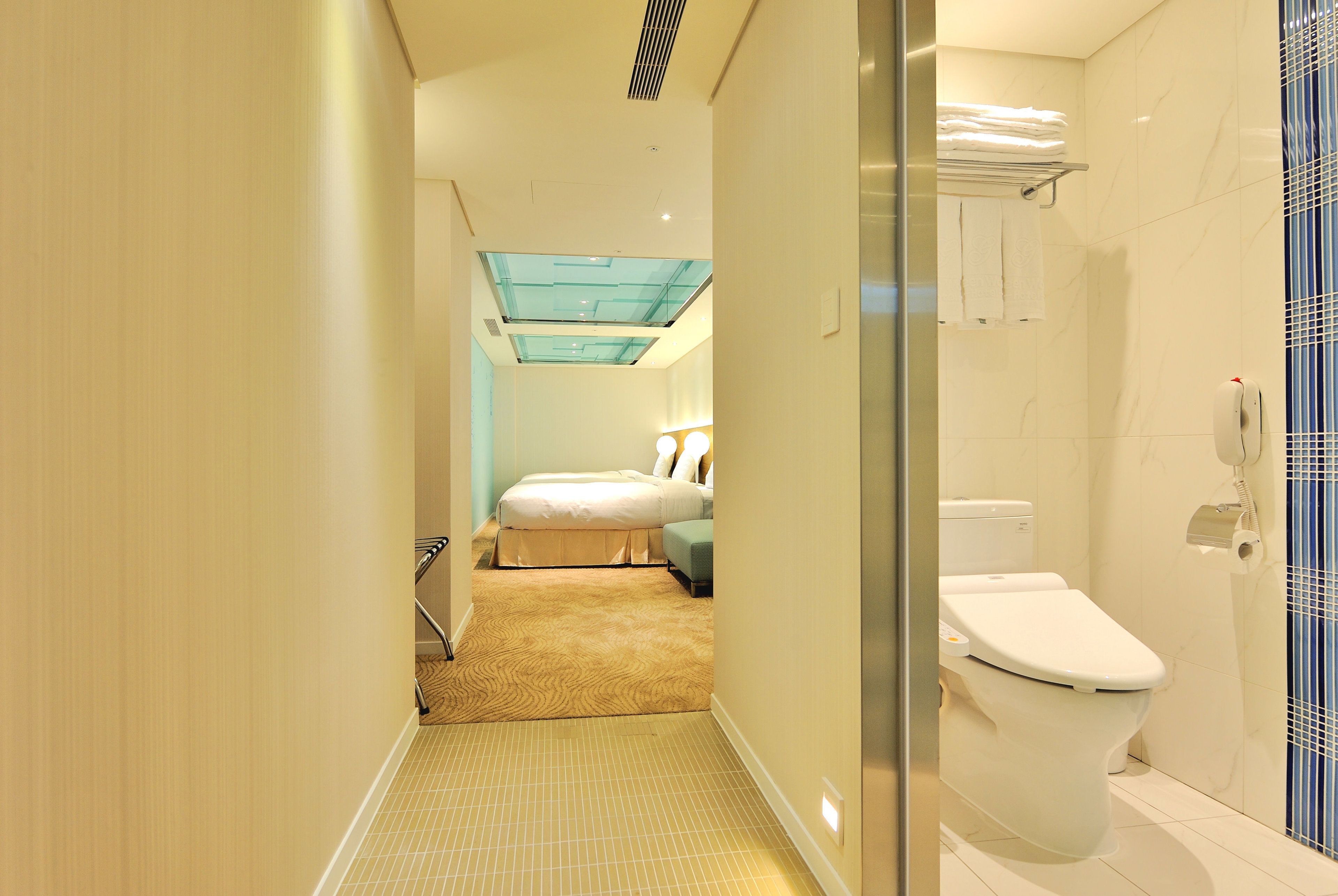 A hotel bathroom with a toilet, a glass shower enclosure, and a bed in the background.