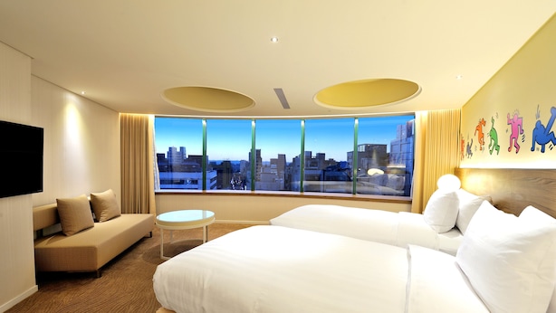 Een hotelkamer met een groot bed, een slaapbank, een klein tafeltje en uitzicht op de stad door grote ramen.