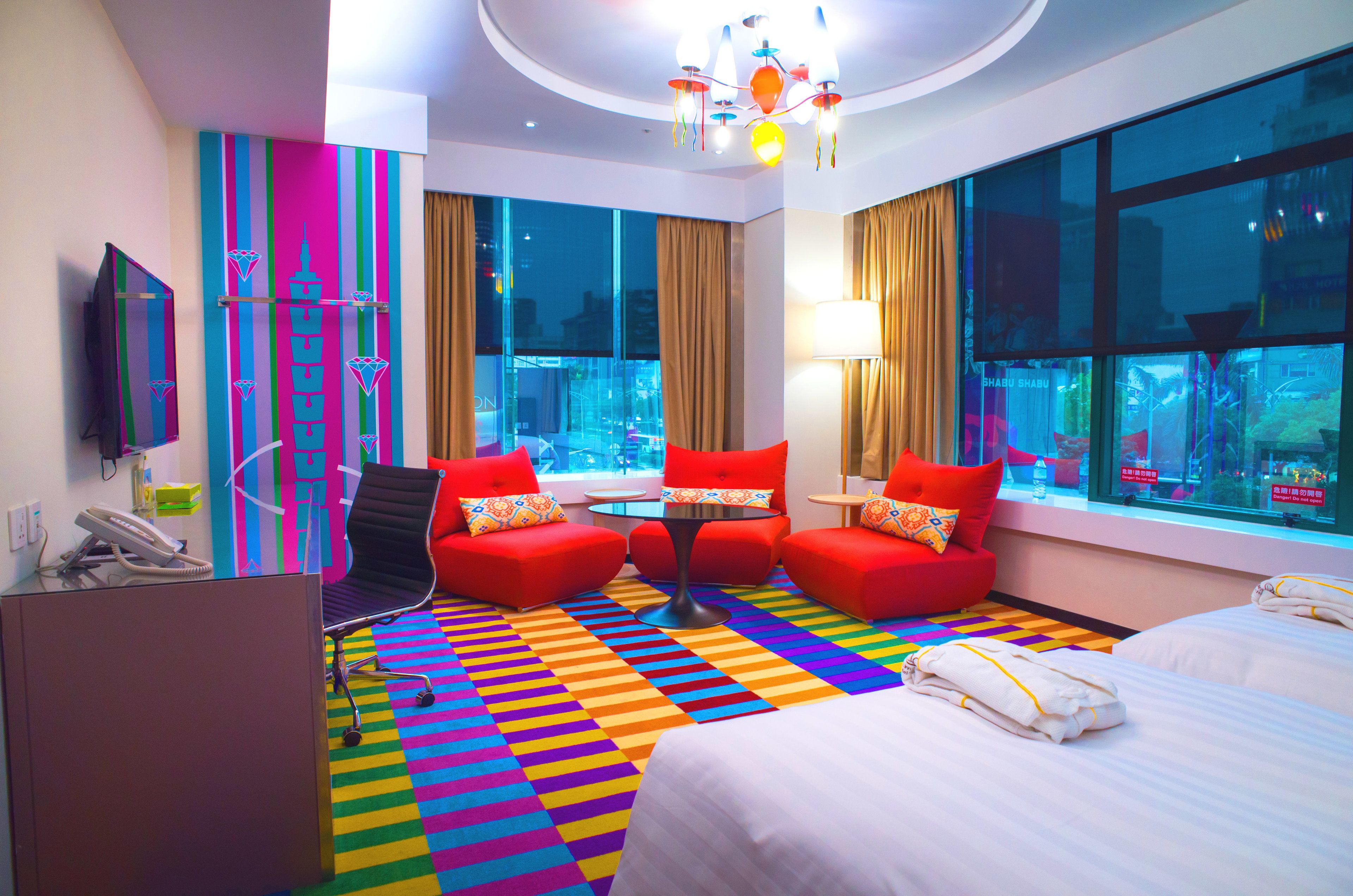 A hotel room with a colorful striped carpet, a bed, two red armchairs, a small round table, and a TV on the left wall.