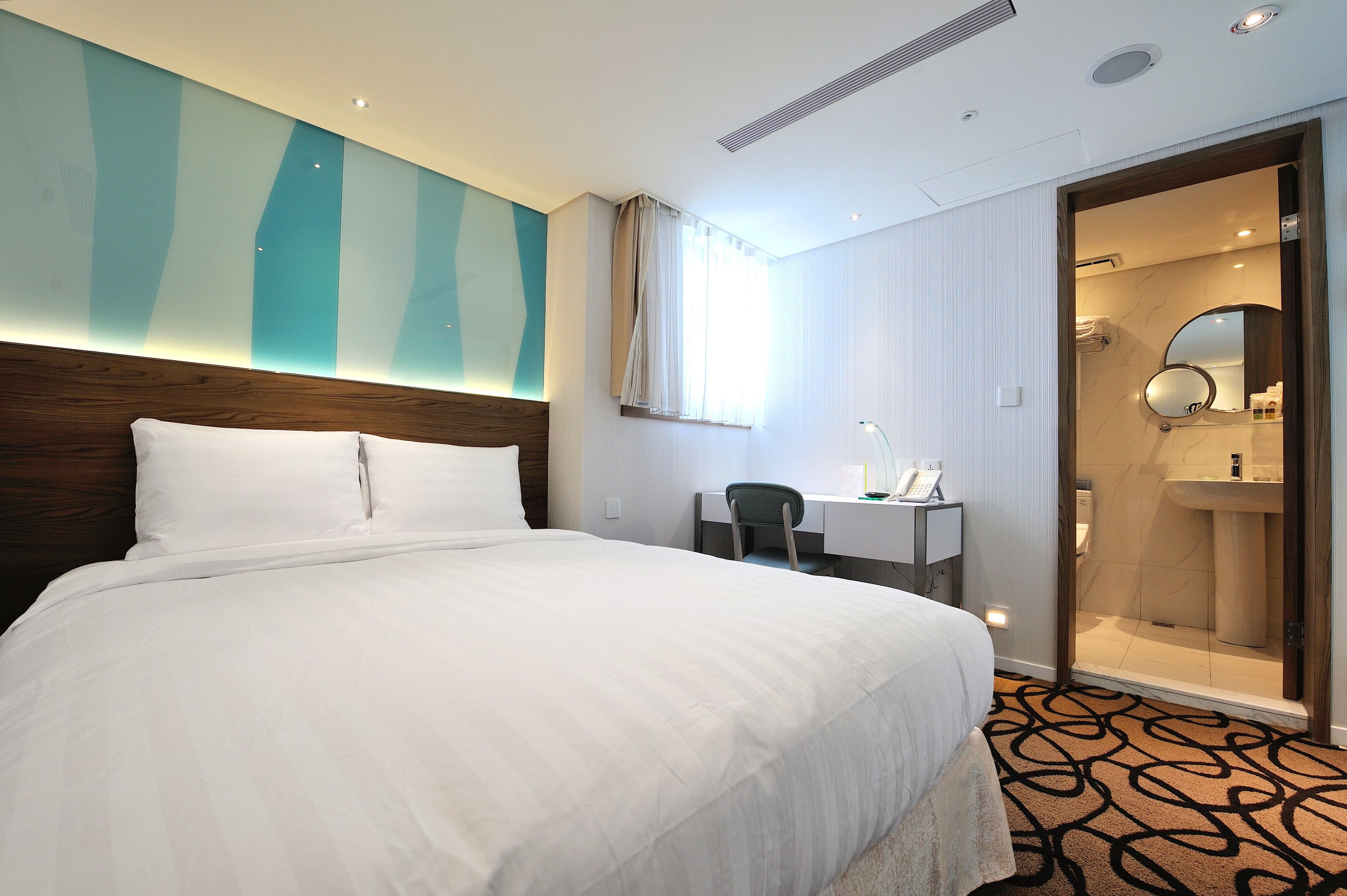 A hotel room with a large bed, a desk, a chair, a bathroom with a mirror, and a patterned carpet.