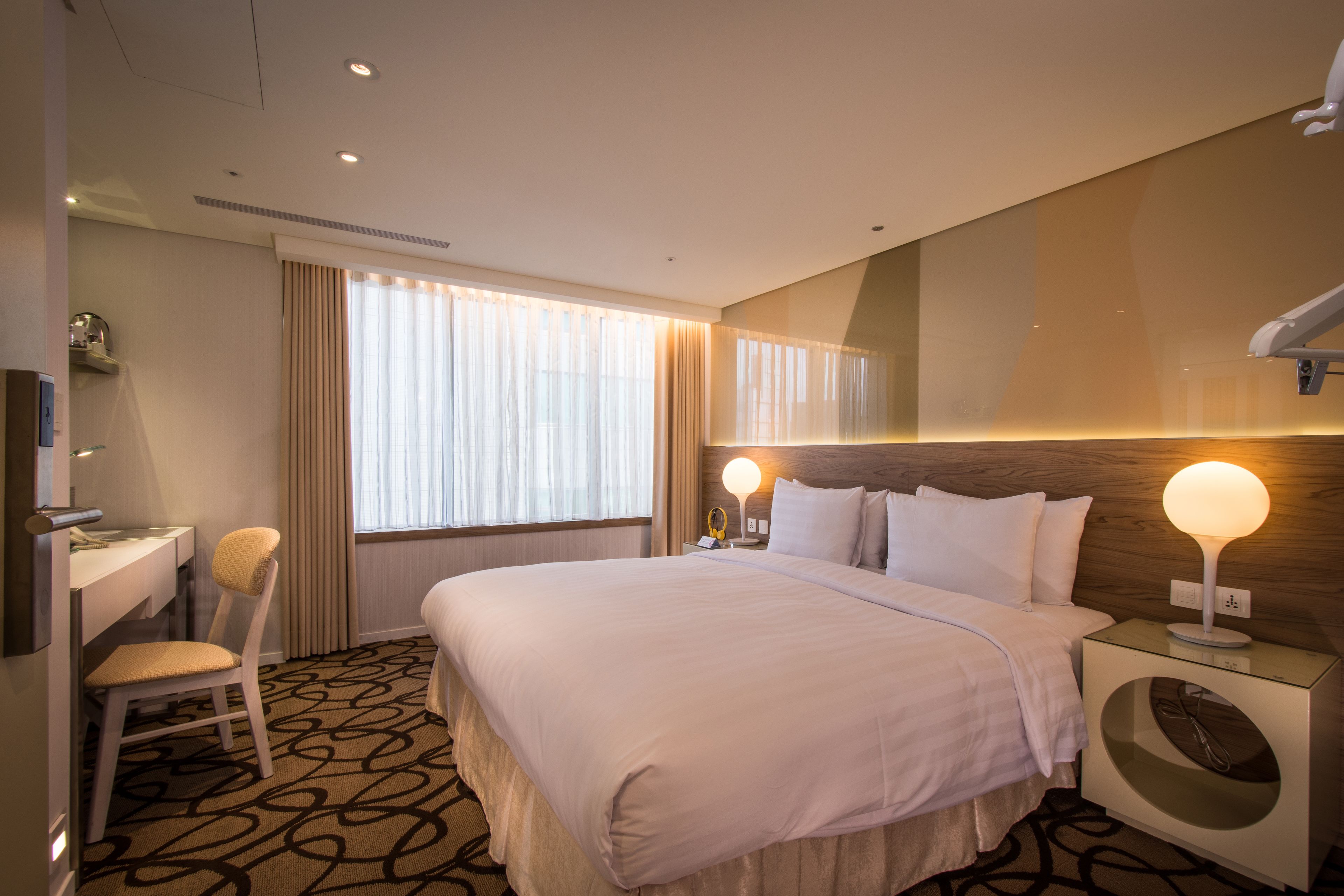 A hotel room with a large bed, a desk with a chair, a vanity, and a wall-mounted lamp.