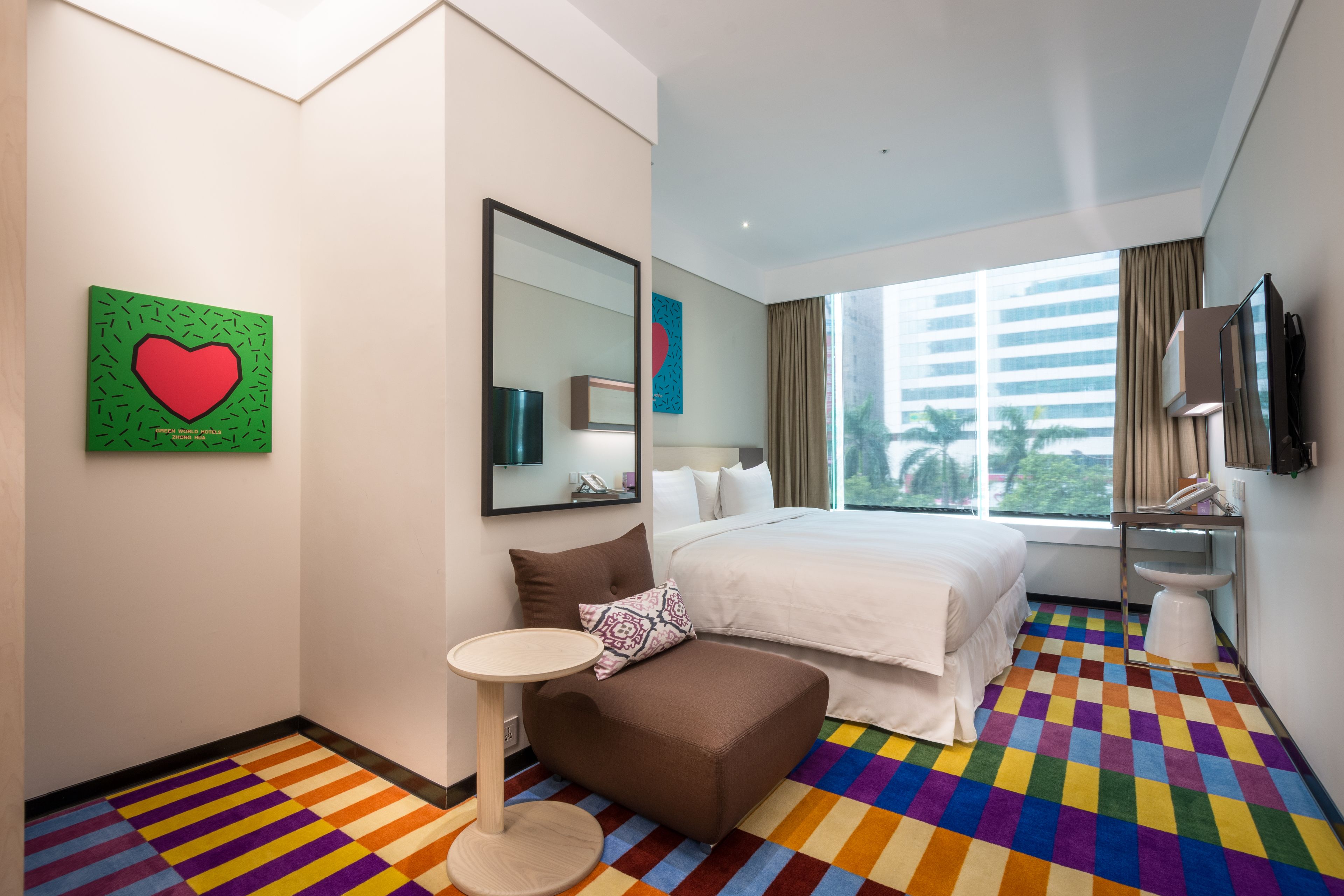 A hotel room with a large bed, a small sofa, a round table, a mirror, and a colorful checkered floor.