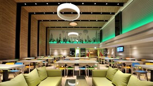 Een moderne eetruimte met meerdere tafels en stoelen, een centrale zithoek met banken, en een groot scherm dat iets toont.