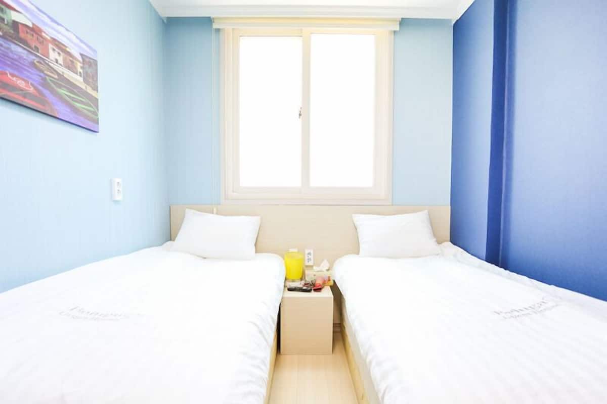 A room with two single beds, a window with blinds, a painting on the wall, and a small table with a bottle and a glass.