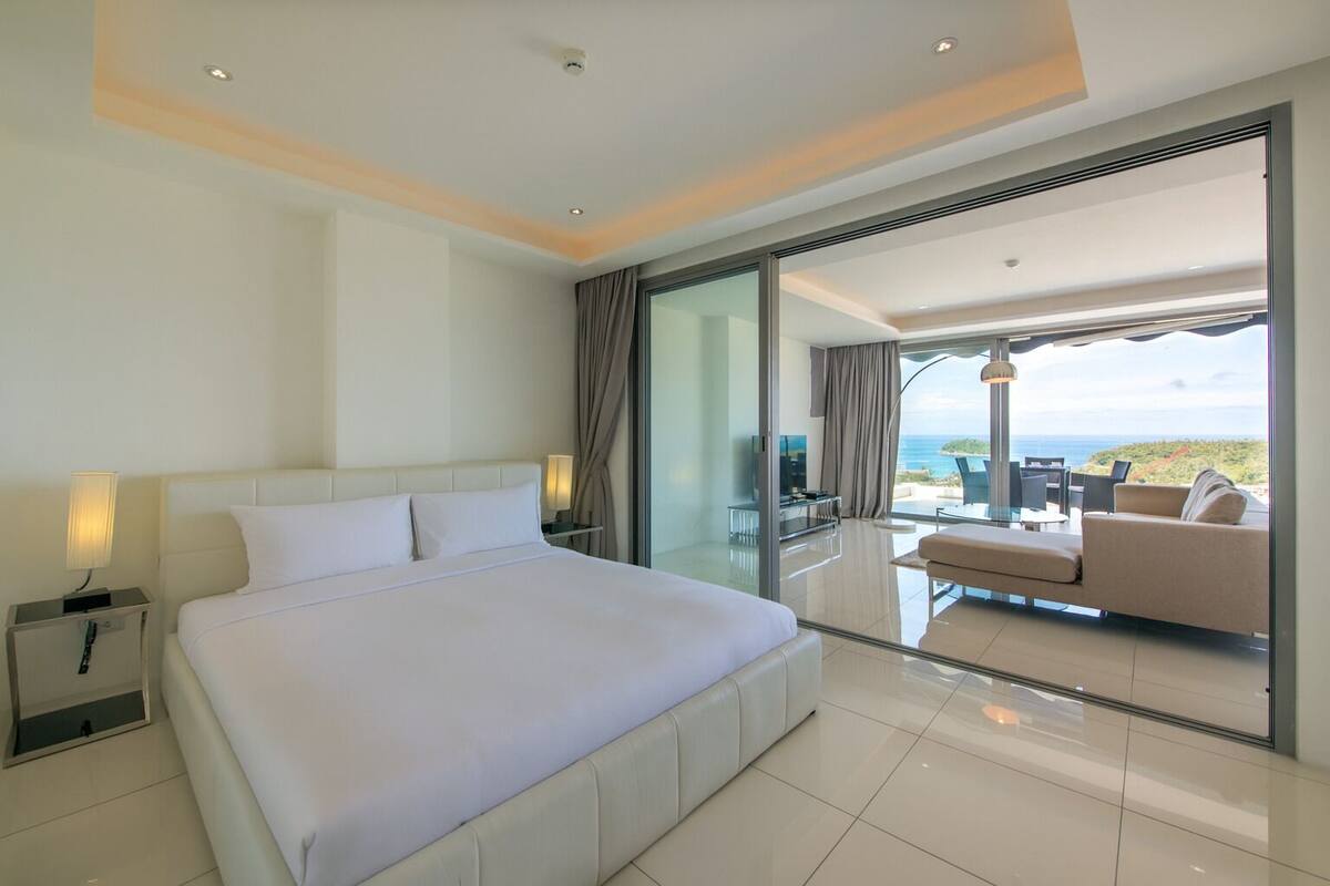 Una habitación de hotel moderna con una cama grande, una puerta de cristal que da a un balcón con vistas al mar, y una zona de estar con una mesa y sillas.