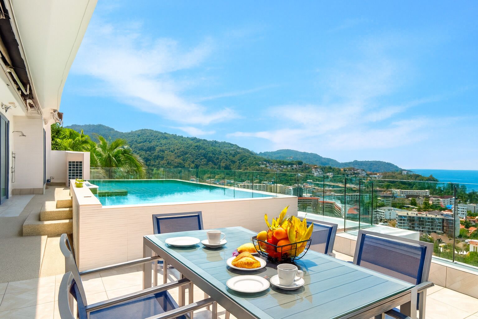 A rooftop terrace with a pool, dining table, and chairs, overlooking a city and hills.