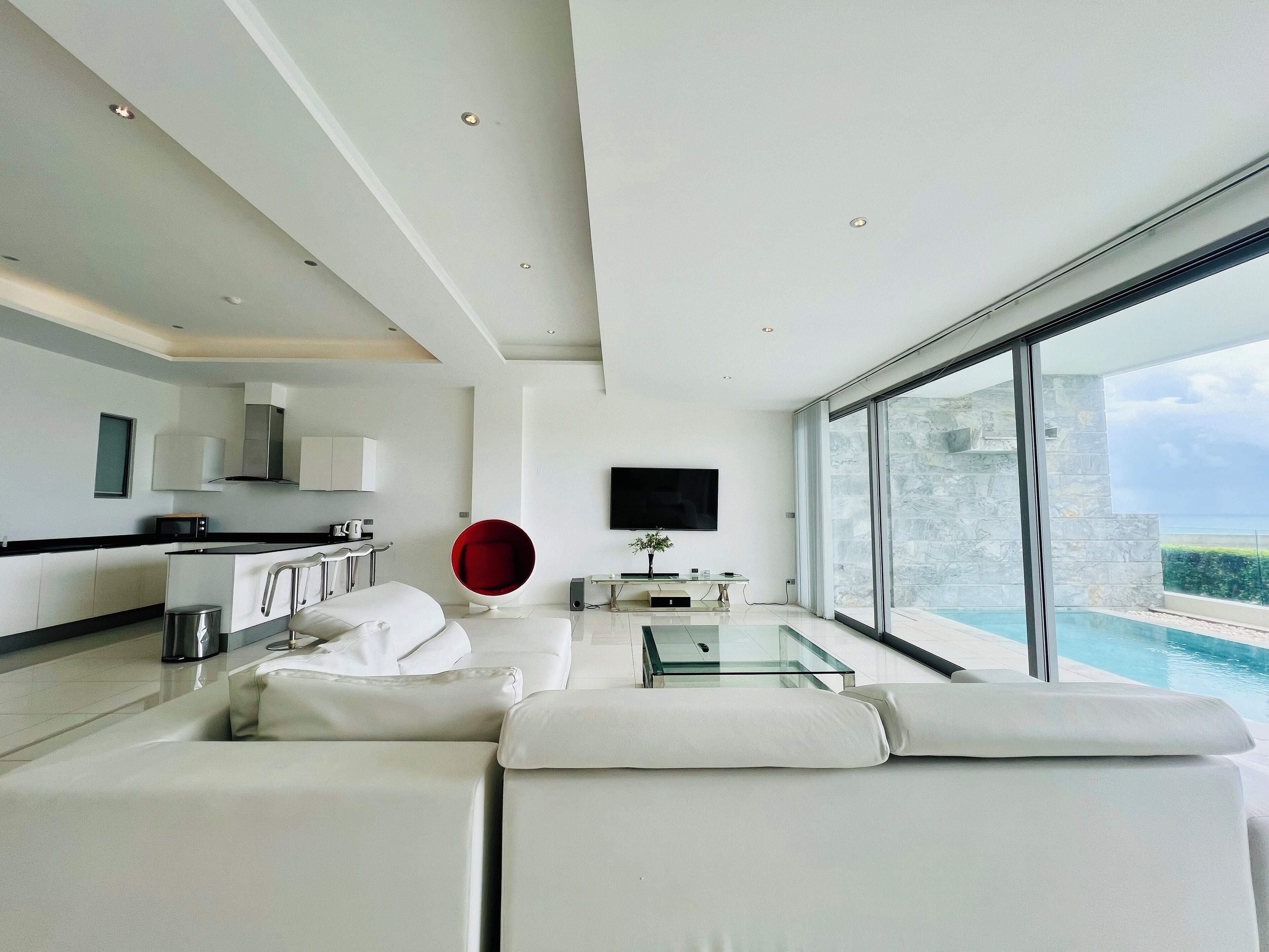 Ein modernes Wohnzimmer mit einem Glastisch, weißen Sofas, einem roten Sessel, einem Flachbildfernseher und Blick auf einen Swimmingpool.