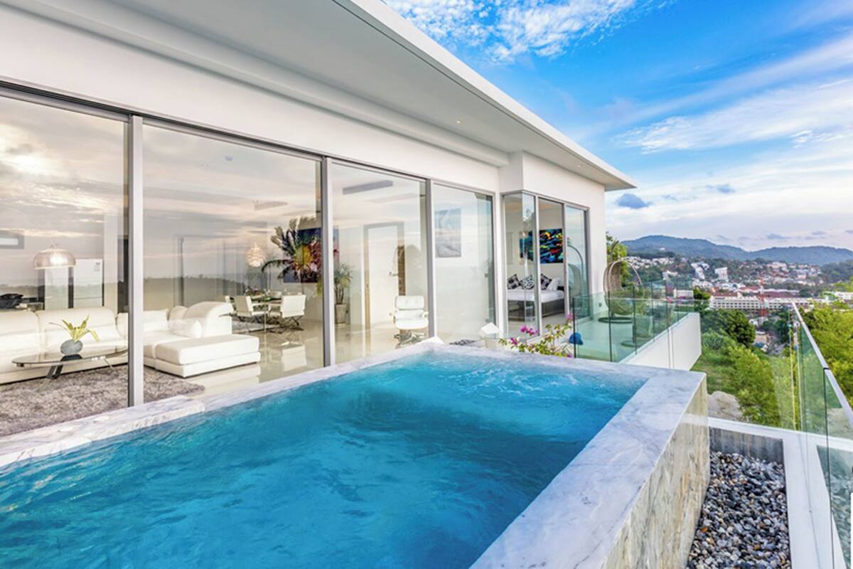 Una casa moderna con piscina infinita envuelta en vidrio, muebles blancos y vistas a la ciudad.
