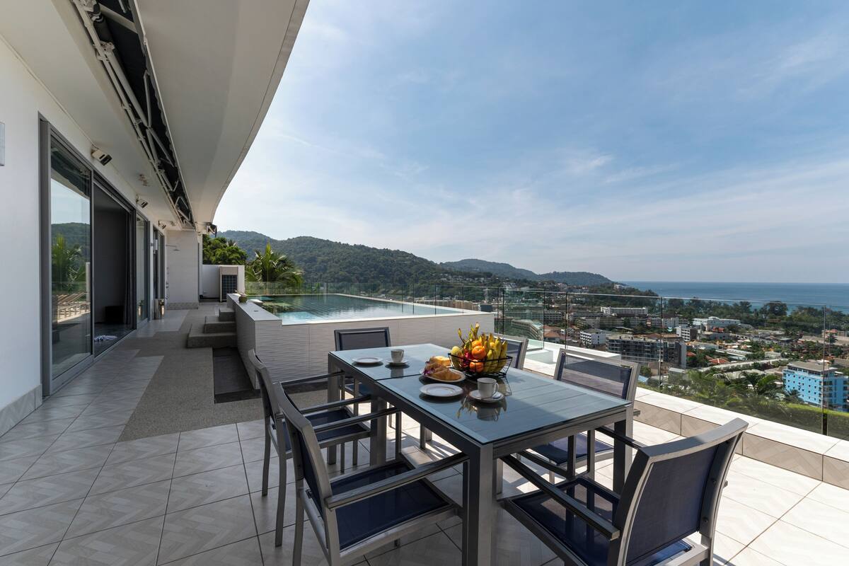 Un área de comedor al aire libre, moderna, con una mesa y sillas de vidrio, con vistas a un paisaje urbano y colinas.