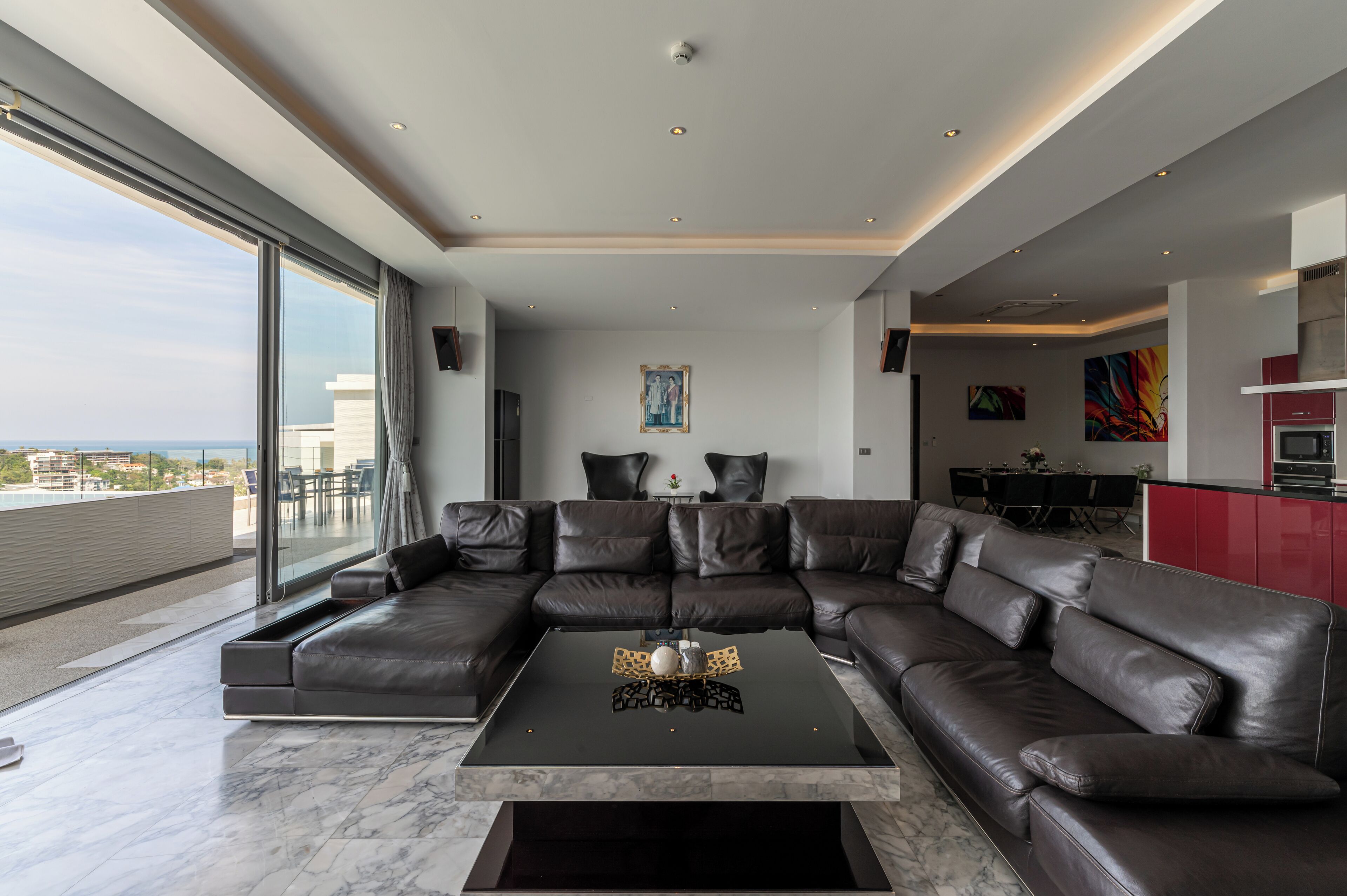 A modern living room with a large sectional sofa, a glass wall with a city view, and a kitchen area with red cabinets.