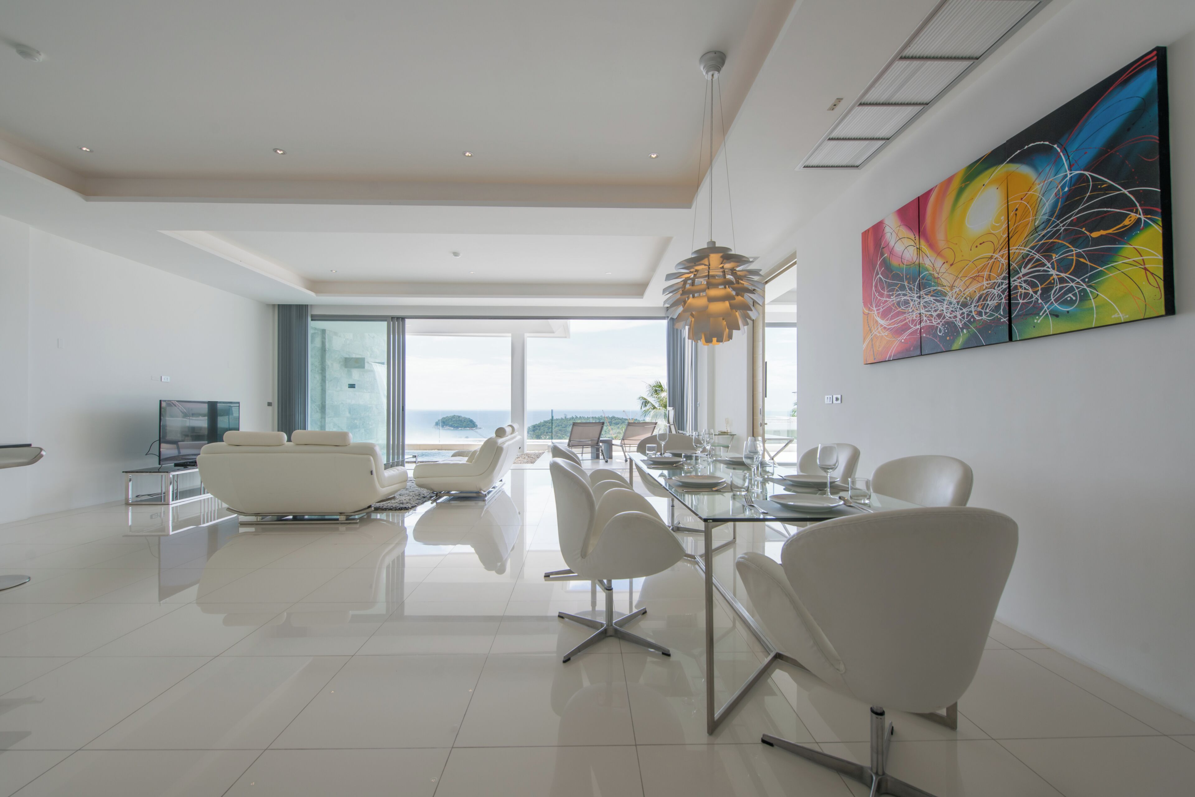 A modern, spacious living area with a dining table, white chairs, and a large window offering a view of the ocean.