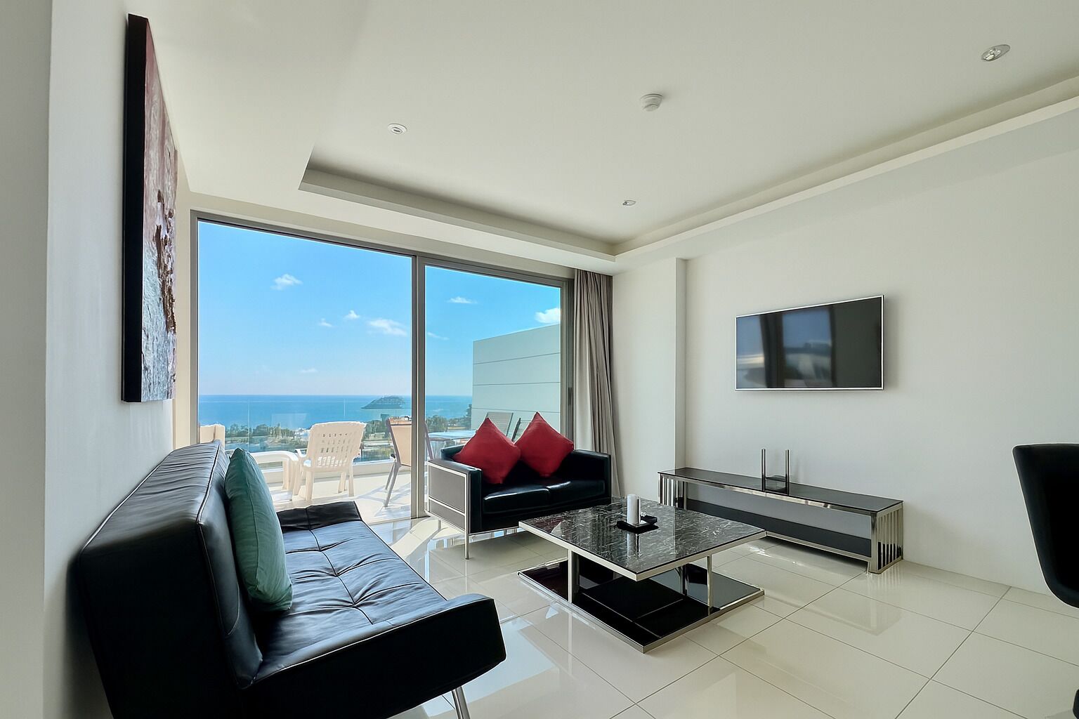 Uma sala de estar moderna com um sofá preto, uma mesa de centro e uma televisão. Há uma grande janela com vista para o mar.