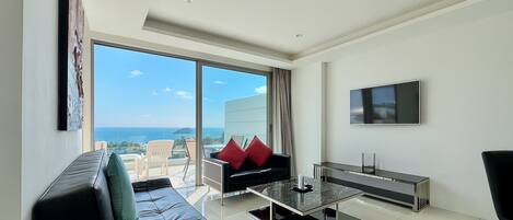 A modern living room with a black sofa, a coffee table, and a TV. There is a large window offering a view of the sea.