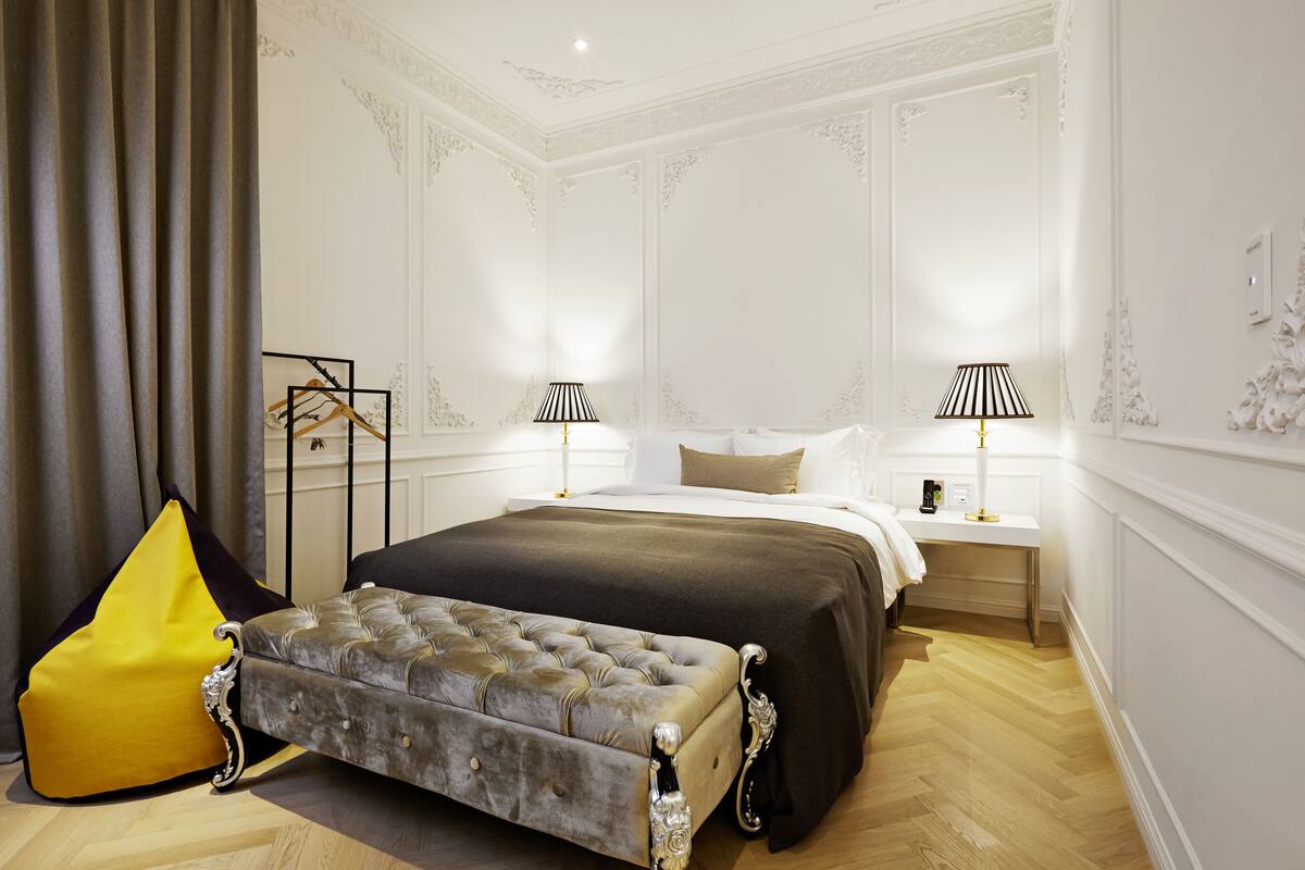 Un dormitorio con una cama grande, dos mesitas de noche con lámparas, un perchero y un banco tapizado.