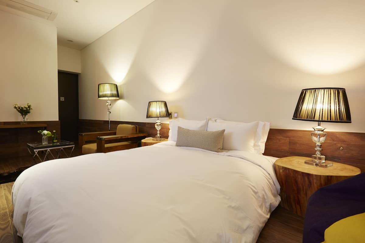 Una habitación de hotel con una cama grande, dos mesitas de noche con lámparas, una pequeña mesa con un florero y un banco de madera.