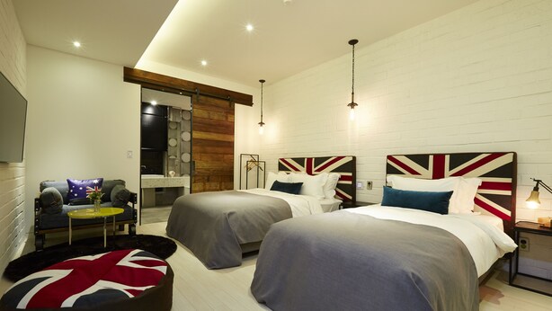 Habitación de hotel moderna con dos camas, un sofá, una mesa de centro y un otoman circular con diseño de la bandera británica.