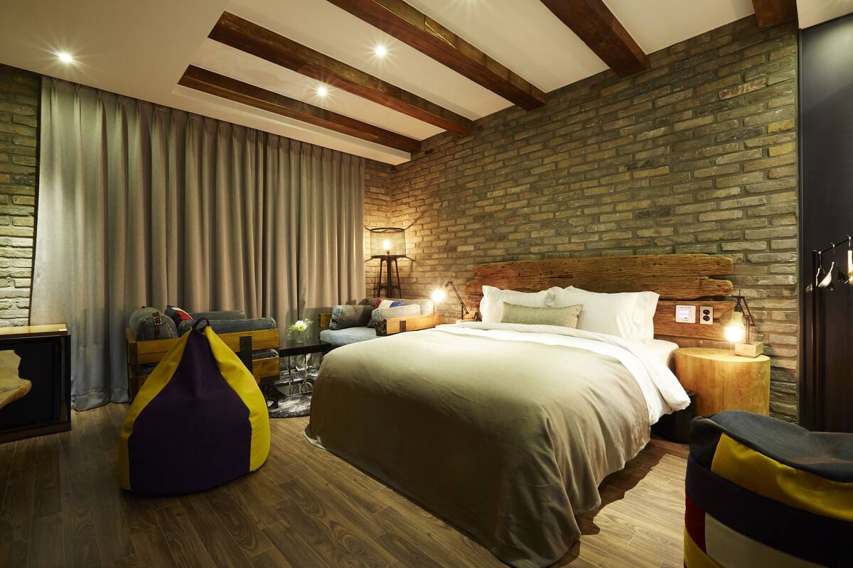 Un dormitorio con una cama grande, vigas de madera, pared de ladrillo y una mesita de noche con lámpara.